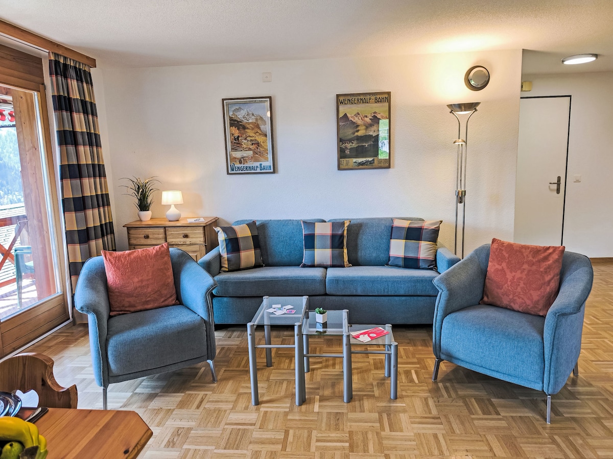 A living area is presented with a comfortable sofa and two armchairs, featuring colorful cushions. A small coffee table sits in the center, complemented by a wooden sideboard and two framed wall art pieces. Natural light filters in through the balcony doors.