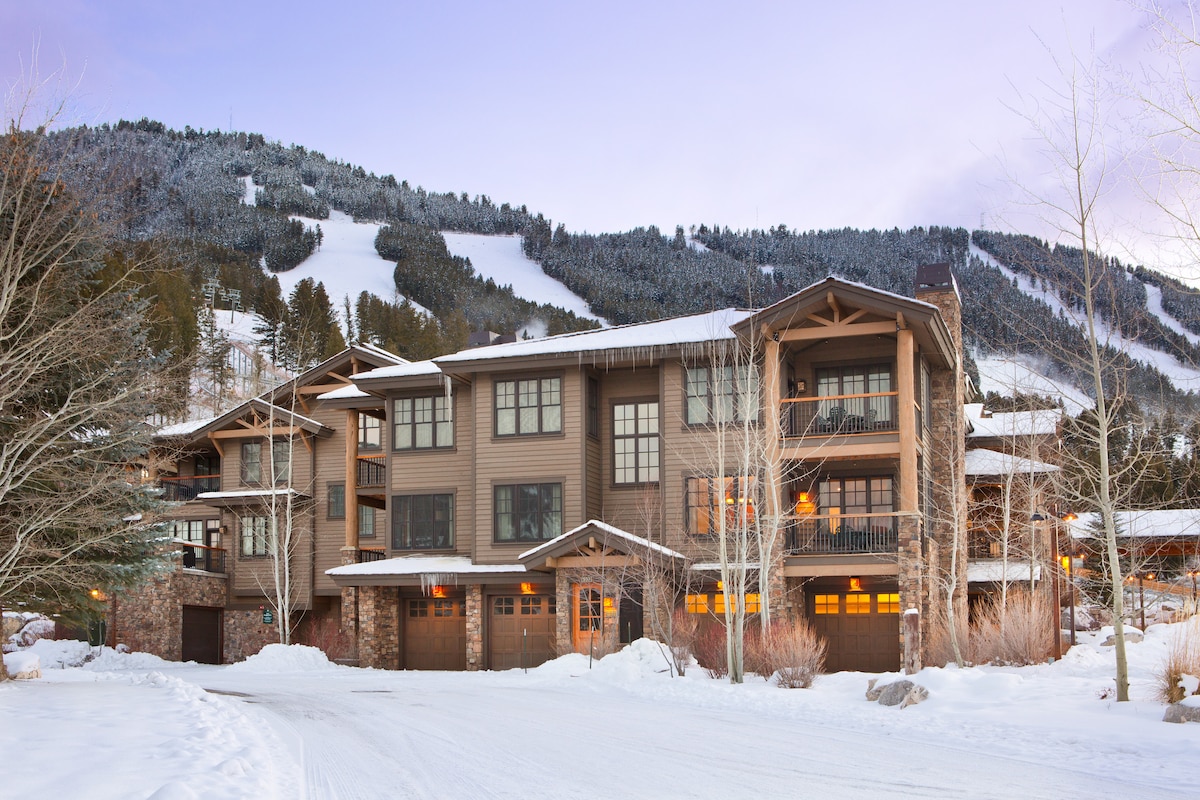 The exterior of a multi-level condo building is shown, framed by snow-covered trees and blanketed ground. The picturesque mountain slopes are visible in the background, indicating proximity to skiing opportunities. Warm light emanates from the windows, creating a welcoming ambiance.