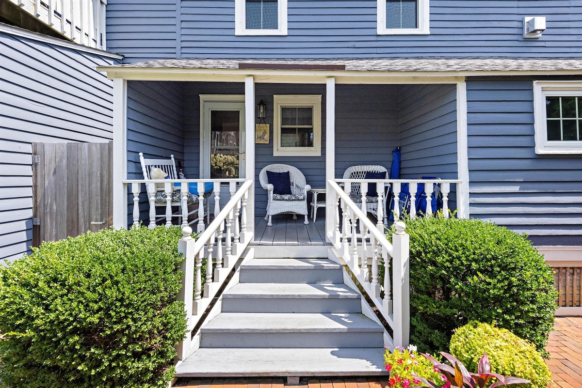 A welcoming porch is adorned with white railings and offers comfortable seating options. Lush green bushes frame the entrance, creating a pleasant view. The shaded area invites relaxation and provides access to the inviting interior of the apartment.