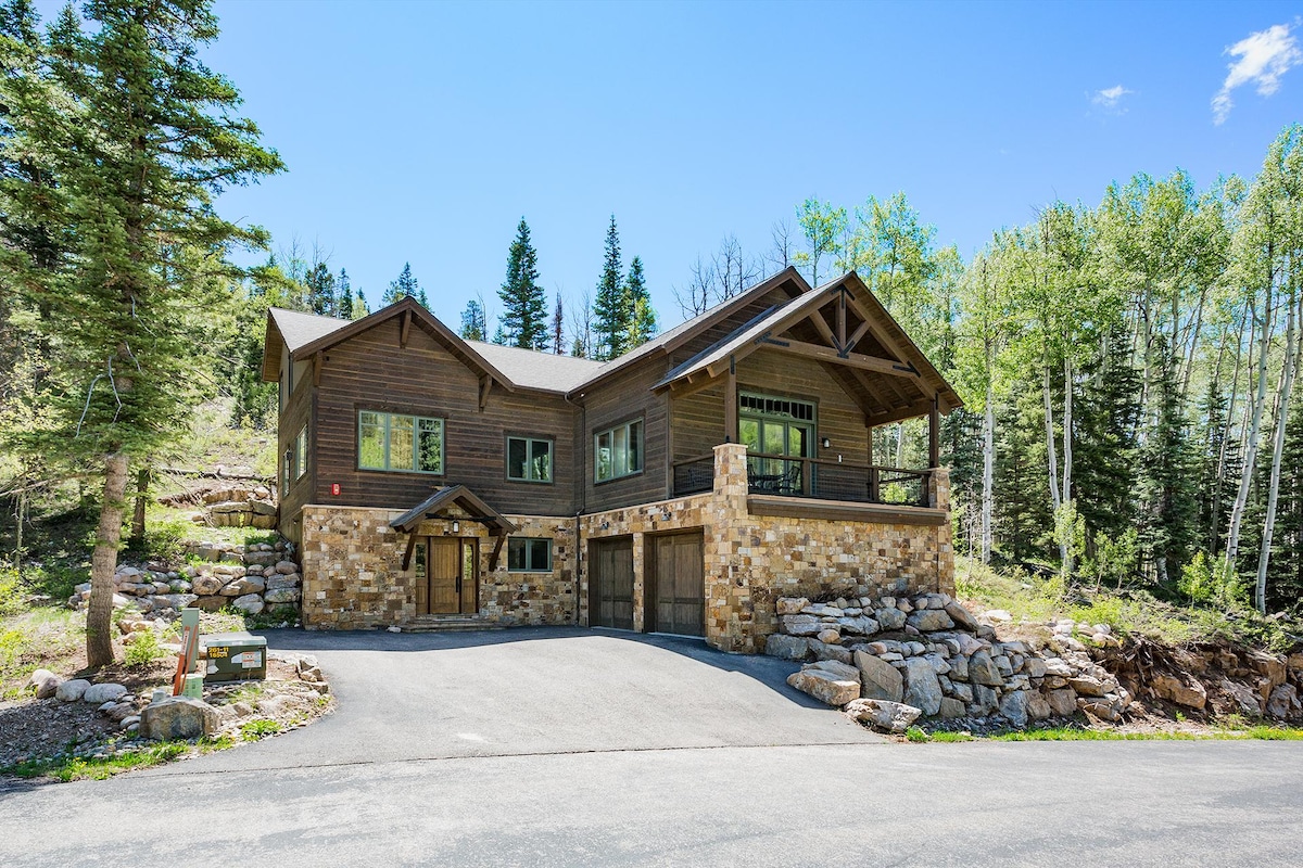 A spacious mountain home is framed by lush greenery, featuring a stone and wood exterior. Large windows offer ample natural light, and a welcoming front porch is visible. The two-car garage is integrated into the design, providing convenient access.