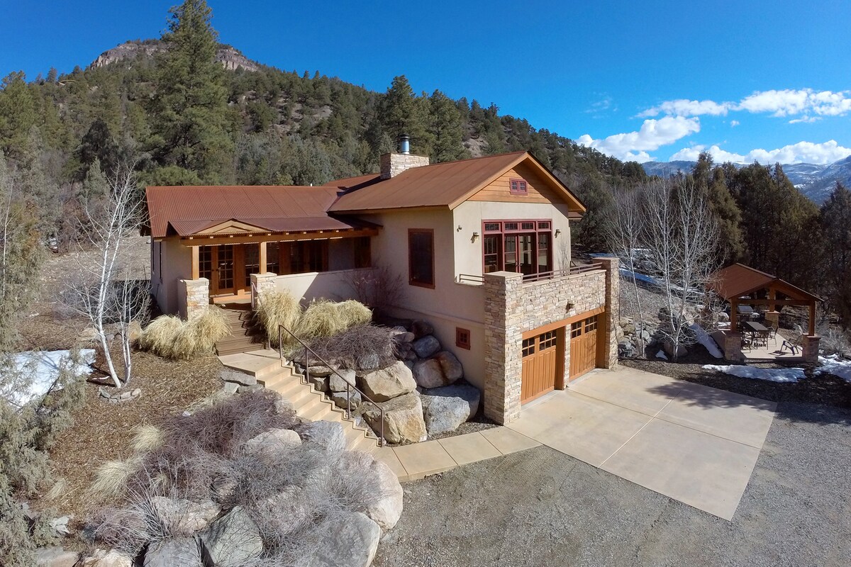 An aerial view showcases the home with a modern design, featuring a stone and wood façade. Surrounding landscaping includes natural rock formations and shrubs, complemented by a driveway leading to the garage. The backdrop features rolling hills and clear blue skies.