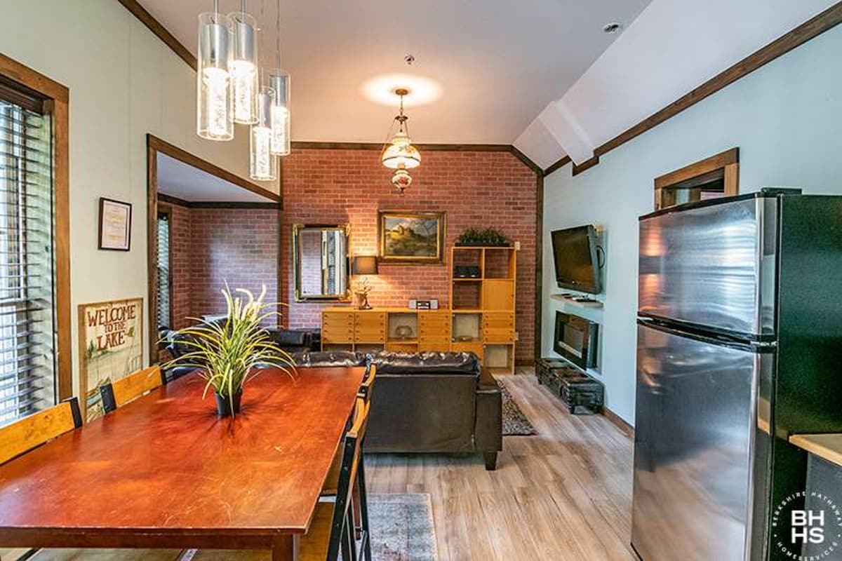 The image features a dining area with a wooden table and chairs, partly illuminated by pendant lights. A brick accent wall is visible, along with a living area containing a dark sofa and entertainment unit. A stainless-steel refrigerator is positioned in the kitchen area.