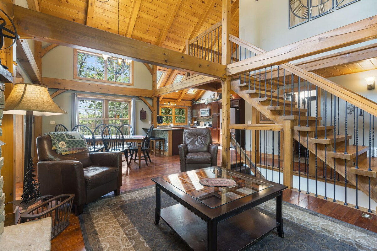 An open living area features a wooden staircase, high ceilings, and natural light streaming through large windows. Cozy seating arrangements include a coffee table and armchairs. The rustic wooden accents are complemented by a view of the dining area and kitchen in the background.