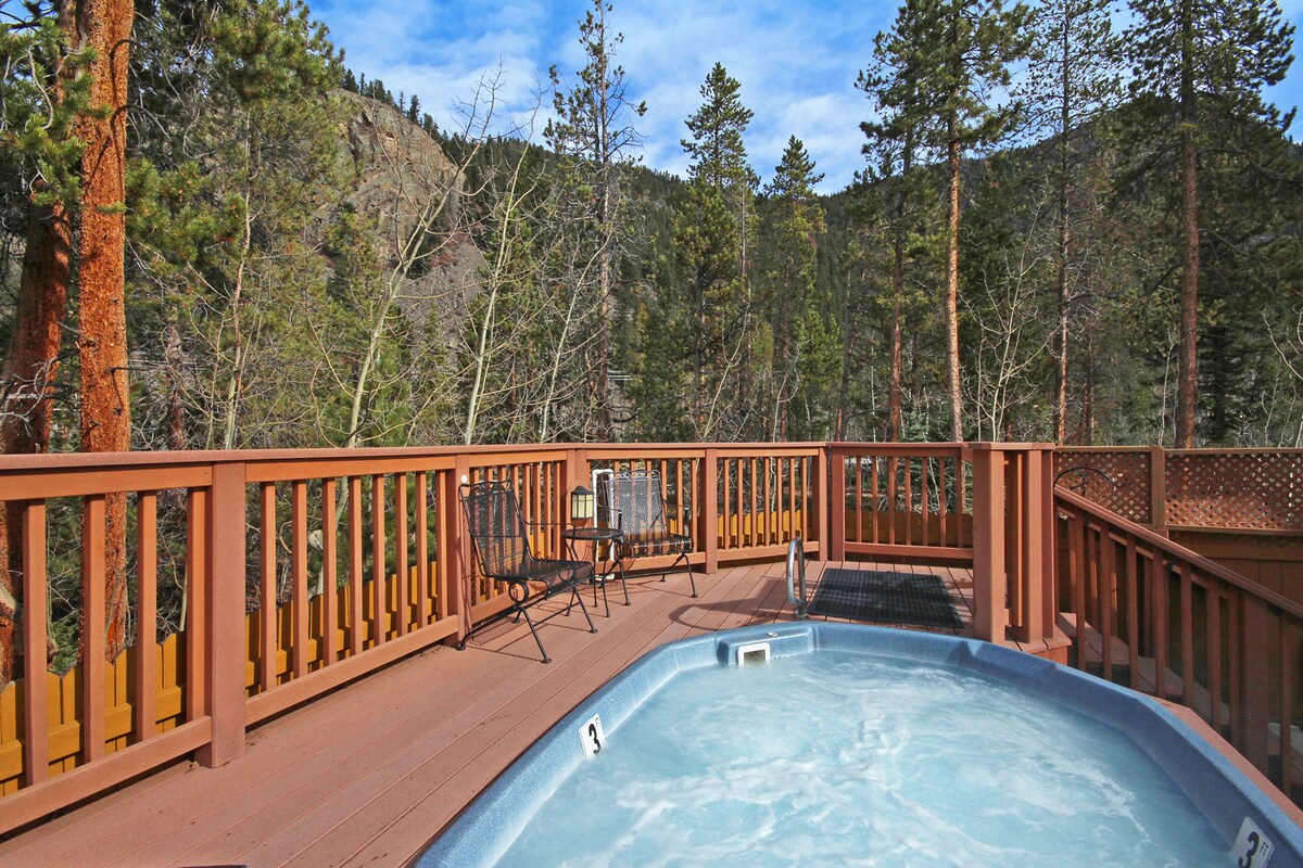 An outdoor shared hot tub is surrounded by wooden decking, offering tranquil views of the forested landscape and distant mountains. Two black chairs and a small table are positioned nearby, inviting relaxation in a serene natural setting.