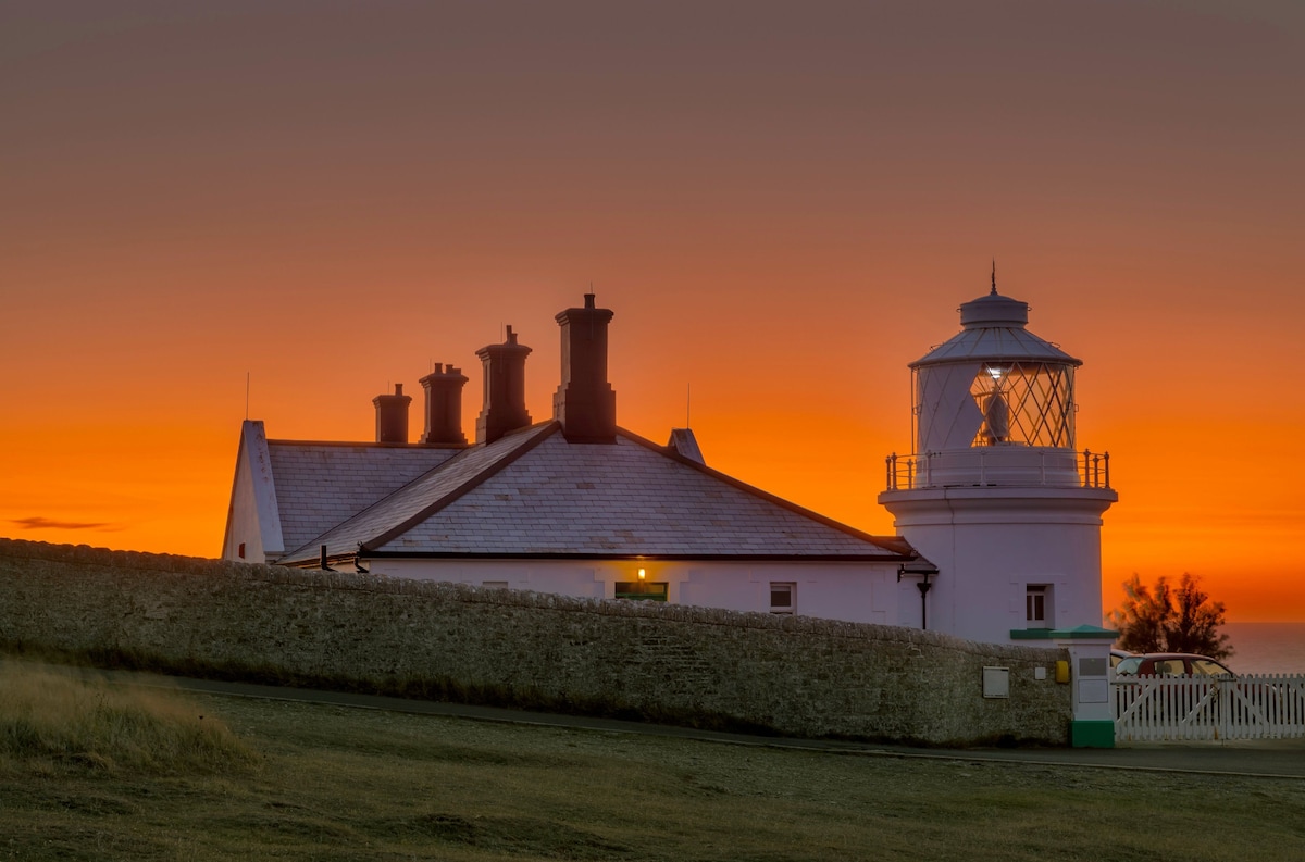 The lighthouse building is shown against a vibrant sunset, with a warm orange and purple sky. The white structure features a lantern tower and a series of chimneys, complemented by a stony perimeter wall that outlines the grounds.