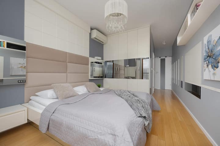 El dormitorio principal tiene una cabecera grande acolchada y un armario empotrado con puertas de espejo. Una colcha gris suave y la iluminación ambiental crean un ambiente de descanso.