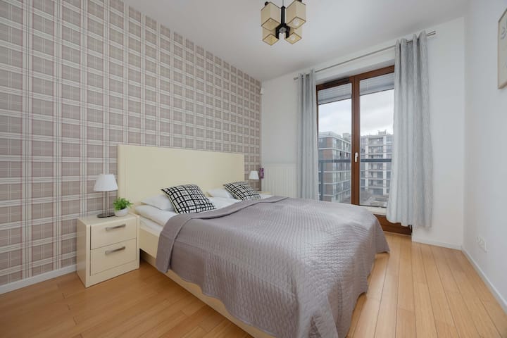 Un dormitorio acogedor cuenta con una cama con una cabecera de color claro y papel tapiz estampado. Las mesitas de noche pequeñas con lámparas se colocan a cada lado para lograr simetría.