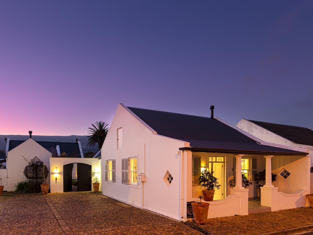 A heritage cottage exterior reveals whitewashed walls and a dark roof, accompanied by decorative diamond-shaped windows. The charming courtyard is paved with cobblestones, and soft lighting illuminates the entrance as dusk approaches, creating a welcoming ambiance.