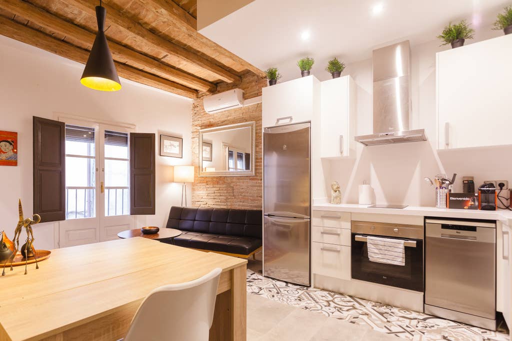 The kitchen area features modern appliances, including a stainless steel refrigerator and an oven with a sleek design. A wooden dining table is complemented by white chairs. Natural light enters through large windows, while original wooden beams and decorative touches contribute to a welcoming ambiance.