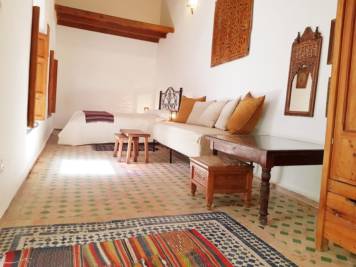 A spacious room features a large sofa with multiple cushions and a low wooden table. A bed is positioned out of view, and natural light filters in through small windows, highlighting a colorful area rug on the tiled floor.