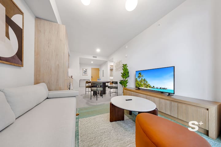 Sala de estar en Miami, Florida, con un sofá de color claro, una mesa de centro redonda blanca, un reposapiés naranja, una alfombra estampada, un televisor montado en la pared sobre un mueble multimedia de madera, arte abstracto en la pared y una vista abierta hacia una mesa de comedor y la cocina.