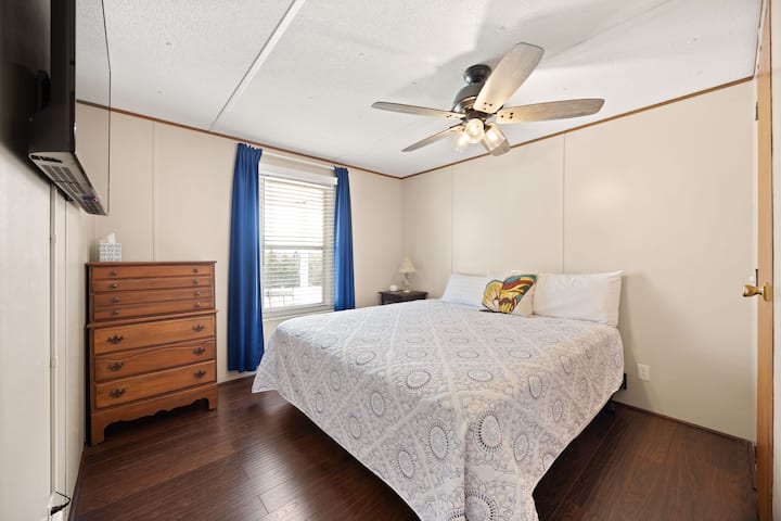 Dormitorio luminoso con cama king, pisos de madera, ventilador de techo y televisor.