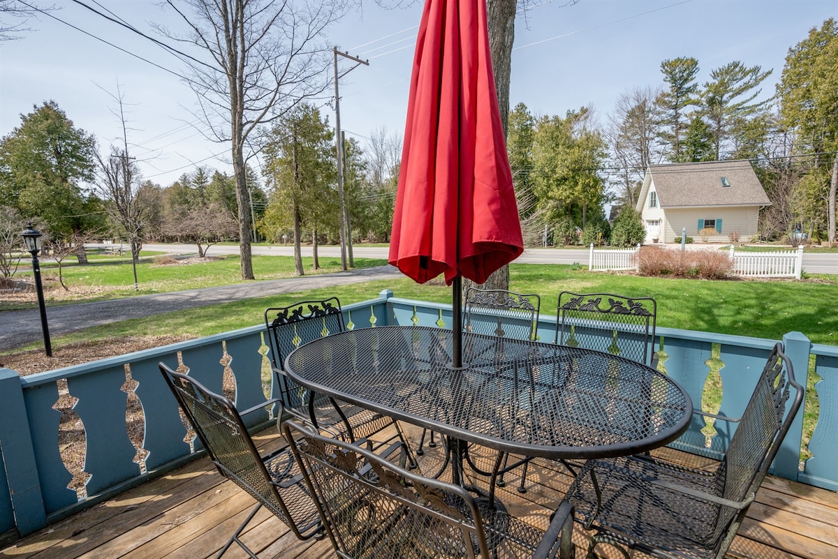 A patio area is shown with a round table surrounded by six black metal chairs. A large red umbrella provides shade. Lush green grass and trees are visible in the background, with a road and home in the distance.