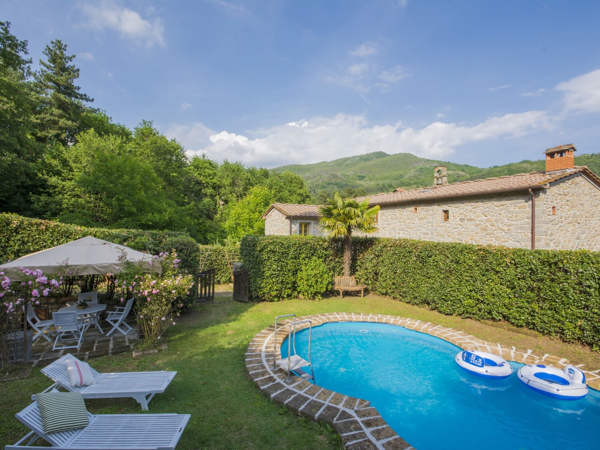 A kidney-shaped swimming pool with bright blue water is surrounded by lush greenery and a small patio area featuring a gazebo and outdoor seating. The serene countryside looms in the background, enhancing the tranquil atmosphere of the well-maintained garden.