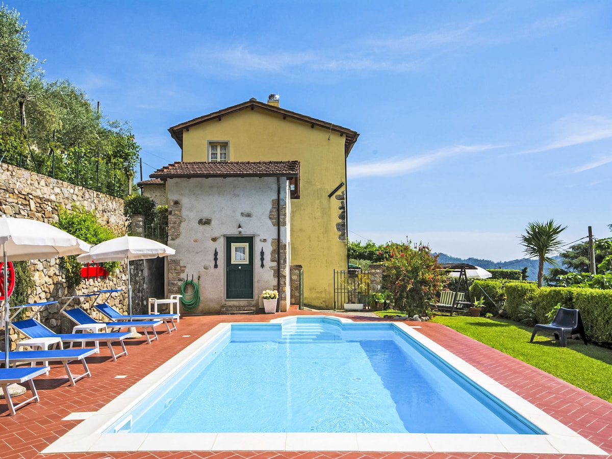 A two-story house with yellow exterior is set beside a clear swimming pool surrounded by terracotta tiles. Lounge chairs are arranged neatly by the poolside, and greenery can be seen in the background, enhancing the serene outdoor setting.