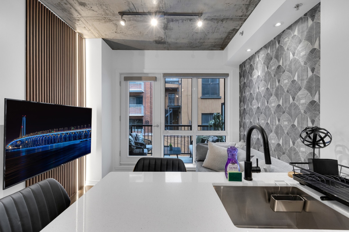 A modern kitchen is visible with a sleek countertop and a sink. A cozy seating area is seen beyond a large window, featuring natural light and a view of surrounding buildings. A television hangs on the wall, showcasing a vibrant image.