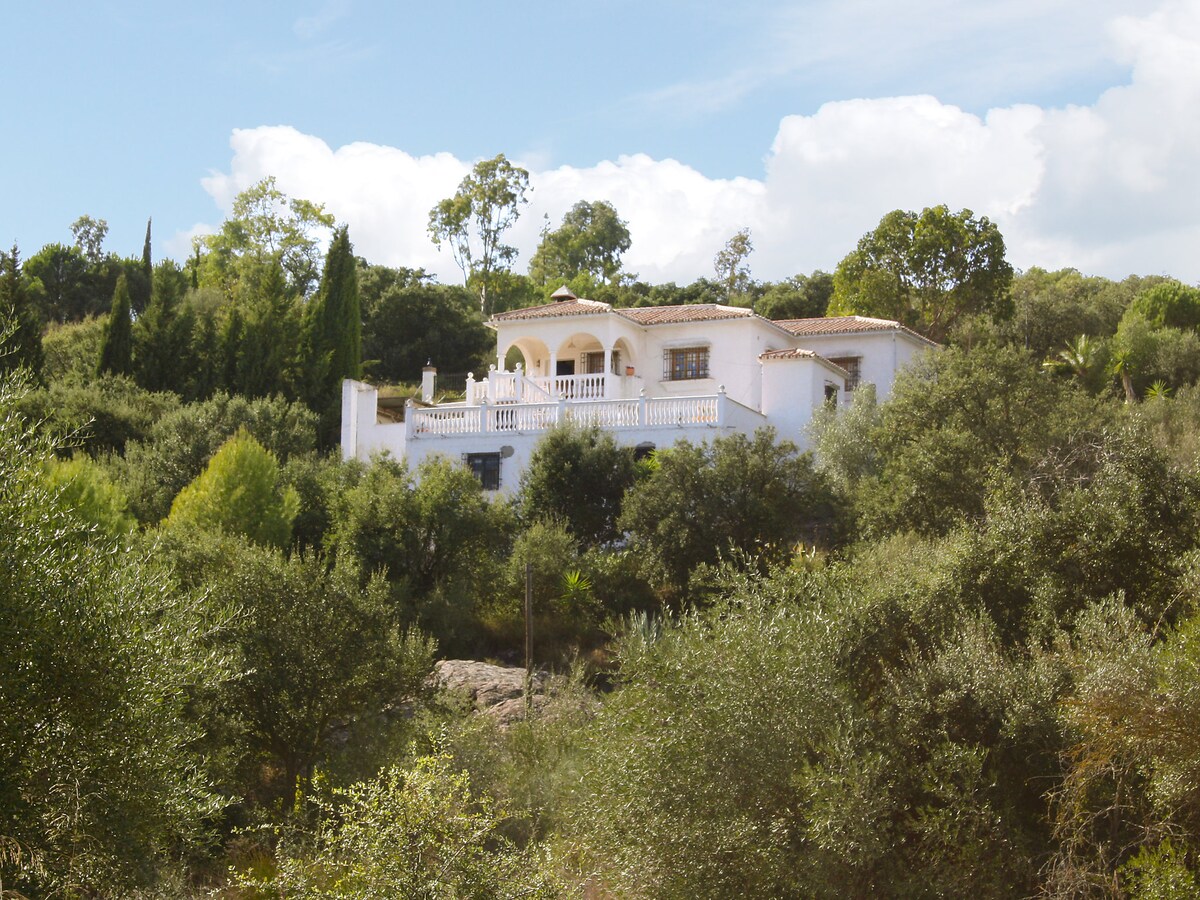 The white two-story house is nestled among lush greenery, surrounded by trees and shrubs. A spacious terrace with decorative railings is visible on the upper level, providing a scenic view of the natural landscape.