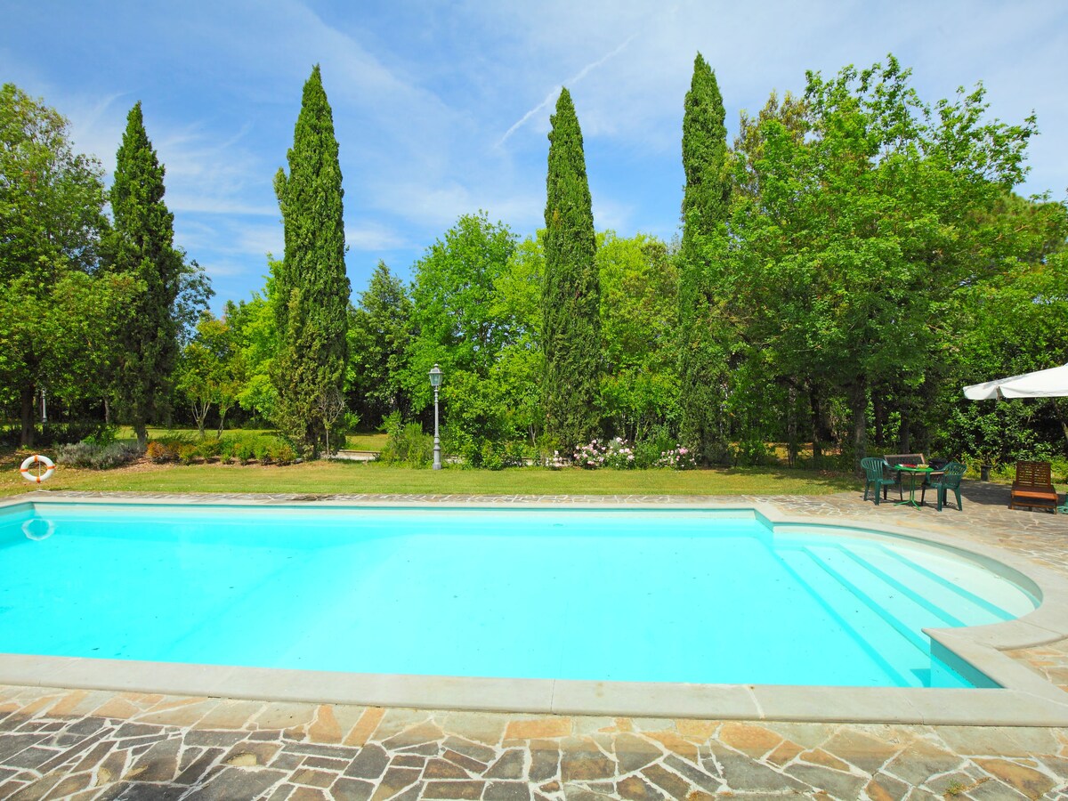 A clear blue swimming pool is surrounded by lush greenery, featuring tall evergreen trees and well-maintained shrubs. A stone patio borders the pool, providing space for lounge chairs and outdoor seating. The serene atmosphere invites relaxation in a natural setting.