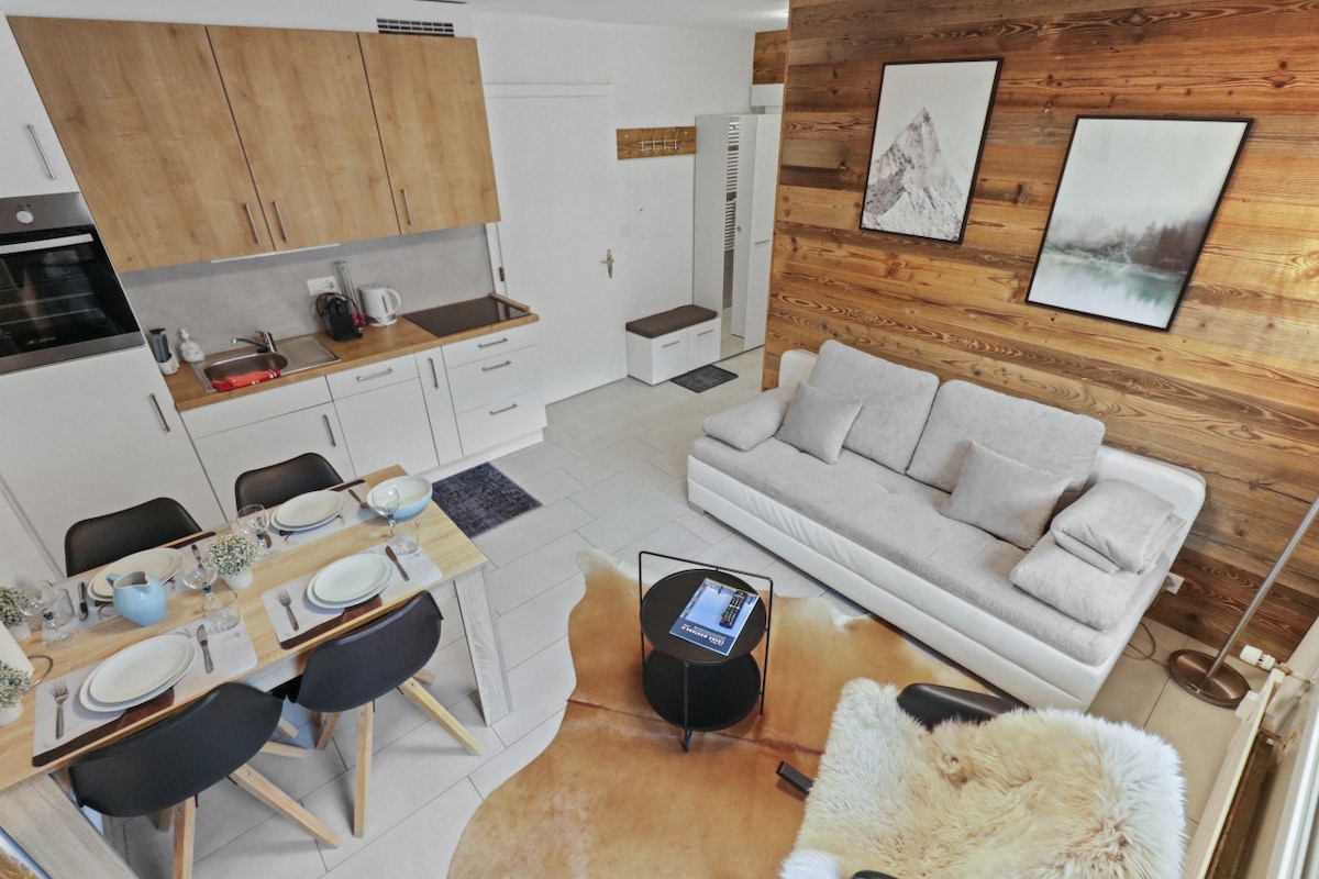 A modern living space is depicted, featuring a light wood dining table set for four with stylish black chairs. A white sofa is positioned beside a wall adorned with wood paneling and framed artwork. The kitchen area is visible in the background, equipped with sleek appliances.
