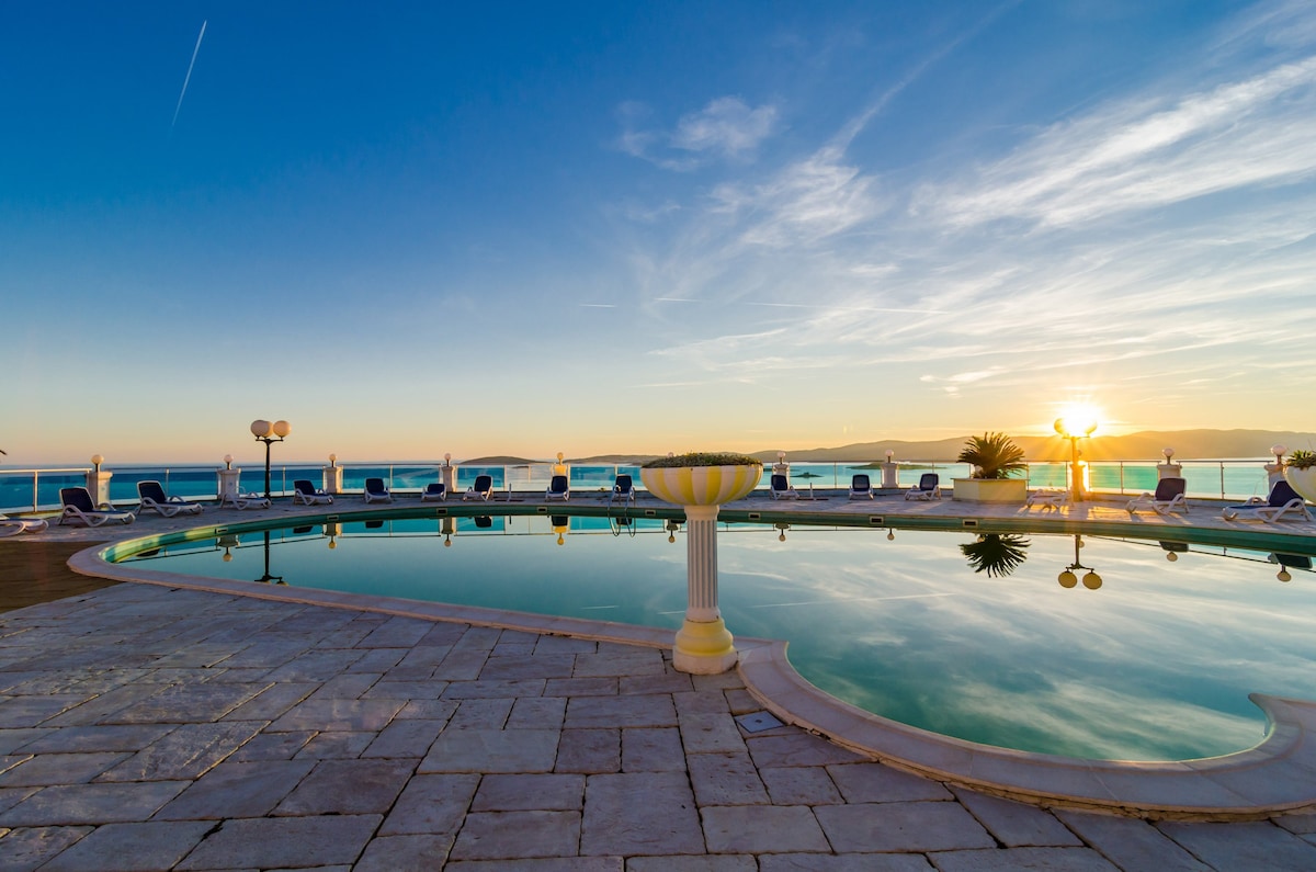 An outdoor pool with a reflective surface is surrounded by sunbeds and umbrellas. The area features stone tiles and column accents, with soft light from the setting sun creating a serene atmosphere. Scenic views of the horizon can be seen beyond the pool.