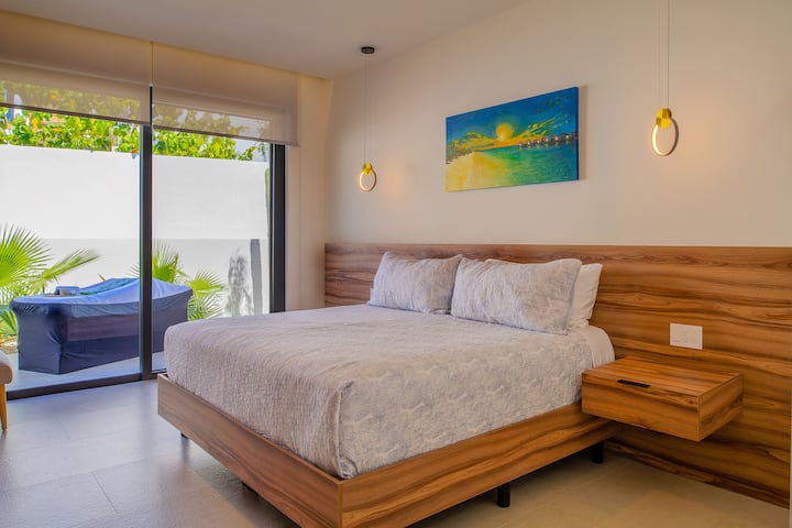 Magpahinga nang payapa sa tahimik mong kuwarto na may mga kaaya‑ayang detalye na gawa sa kahoy, modernong pendant lighting, at mga tanawin ng hardin sa mga bintanang abot sa sahig at kisame.