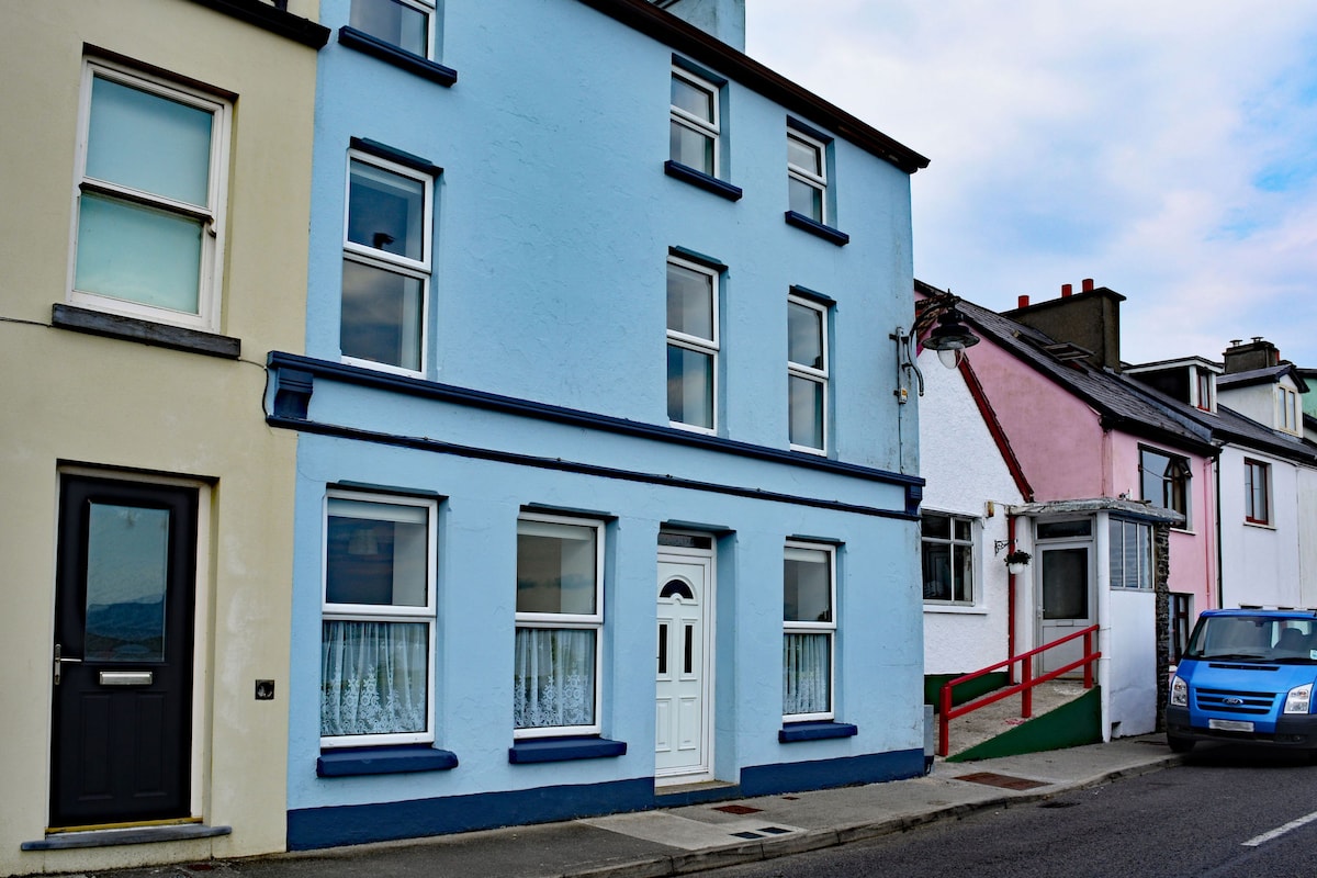 The exterior of a charming multi-level building is displayed, featuring a blue facade with several large windows. A white door is centered at the front, with lace curtains visible through the glass. Adjacent buildings in pastel colors add to the village character.