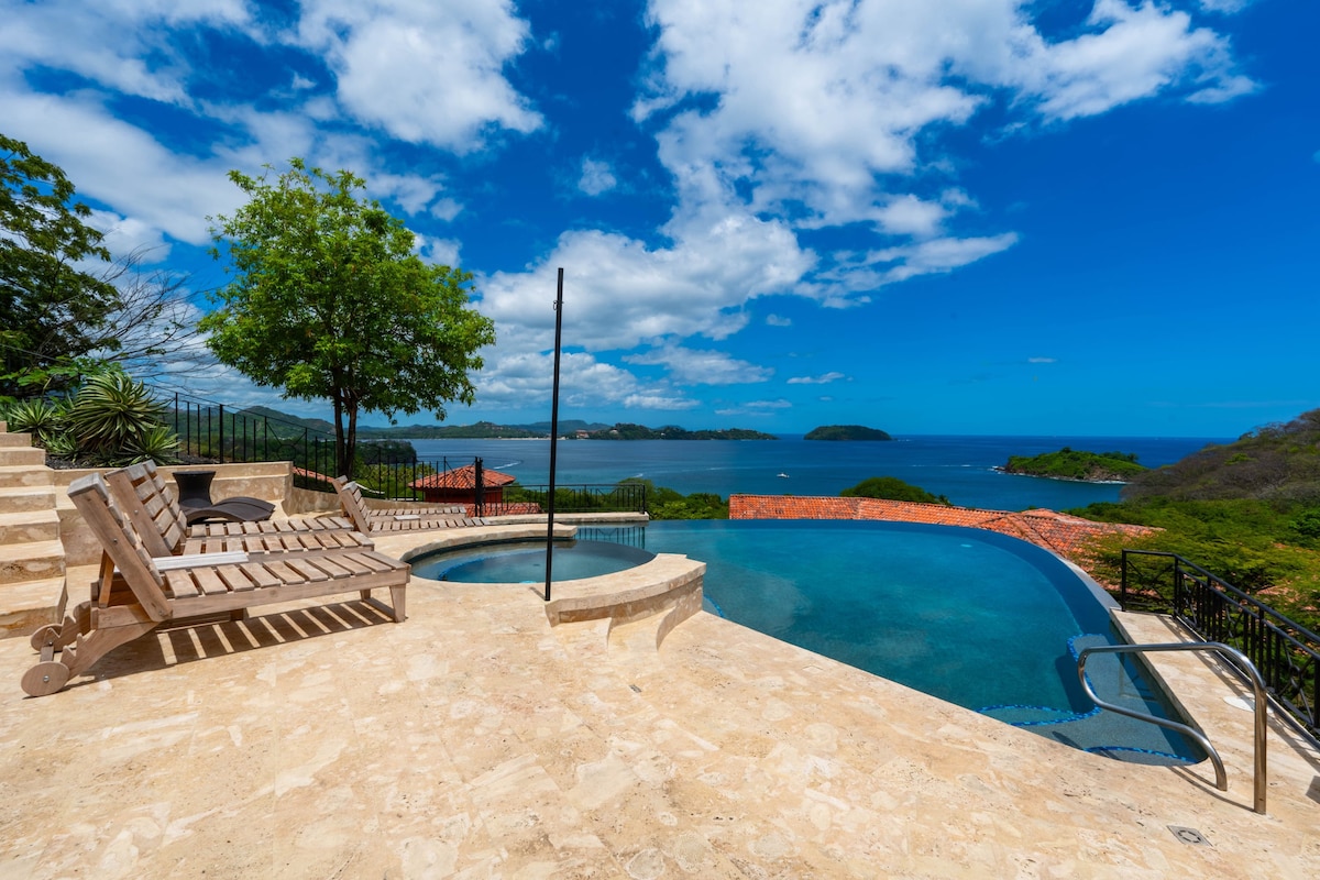 An infinity pool is positioned on a sunlit patio, overlooking the ocean. Comfortable lounge chairs are arranged nearby, and a small hot tub is integrated into the pool area. Lush greenery surrounds the space, complementing the stunning views of the ocean and distant islands.