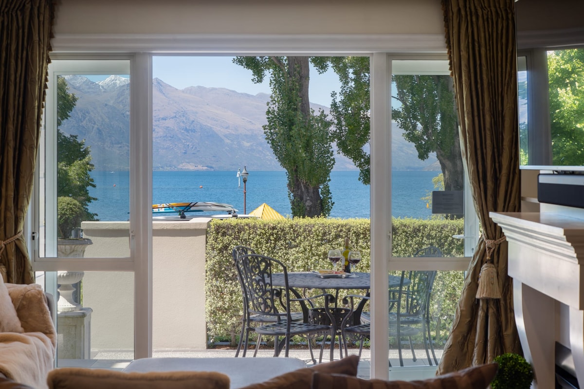A view from the living area frames a serene lakefront scene, featuring outdoor furniture set on a private patio. Lush greenery surrounds the space, while the tranquil waters of Lake Wakatipu and distant mountains create a picturesque backdrop.
