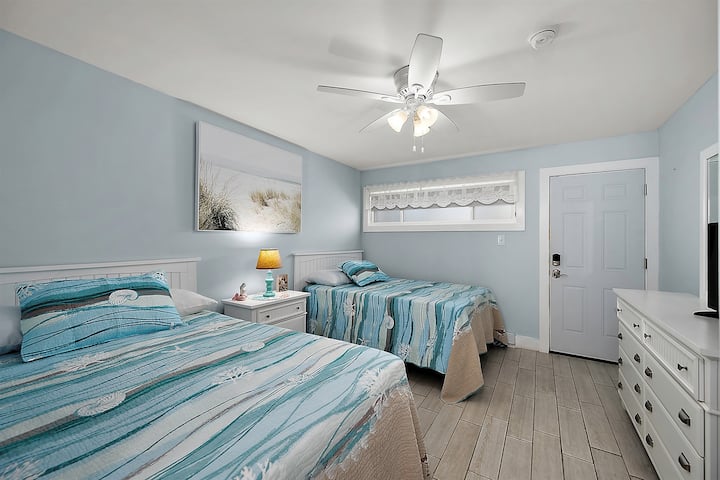 Dormitorio costero con dos camas cómodas, sábanas suaves y ventilador de techo para mantenerte fresco después de un día en el agua.