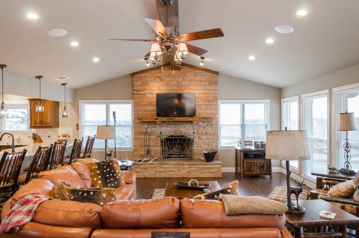 A spacious living room features a warm stone fireplace as the focal point, surrounded by large windows that allow ample natural light. Comfortable seating is arranged in an inviting circular layout, complemented by wooden accents and stylish lighting fixtures.