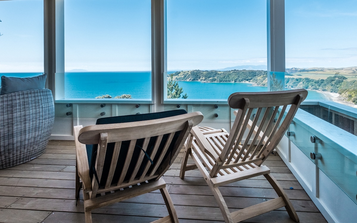 Two wooden recliners are positioned on a balcony overlooking breathtaking ocean views. Floor-to-ceiling windows create an open atmosphere, allowing natural light to fill the space. The tranquil setting invites relaxation while offering panoramic vistas of the water and coastline.