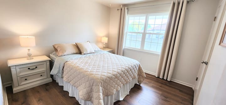 Habitación luminosa y acogedora con cama queen, piso de madera y una ventana grande.