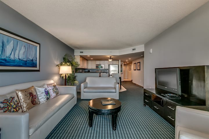 This stylish living area features modern white seating, a dark wood coffee table, and nautical artwork. The open-concept layout flows into a fully equipped kitchen, while striped carpeting and warm lamp lighting create an inviting atmosphere for gues