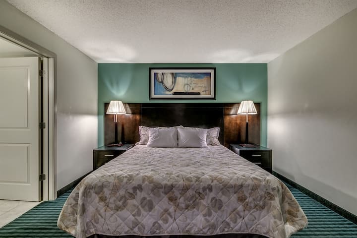 This welcoming bedroom features a large bed with a patterned quilt and white pillows. A dark wood headboard is set against a teal accent wall with framed art, flanked by two nightstands with elegant lamps. Striped carpeting adds a touch of modern sty