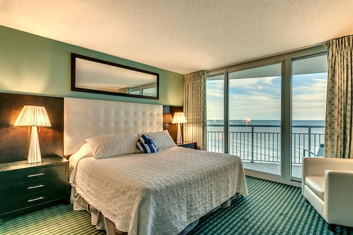 Wake up to breathtaking ocean views in this primary bedroom. It features a king bed with a tufted white headboard, crisp white linens, and a striped accent pillow. Large sliding glass doors open to a private balcony overlooking the beautiful coastlin