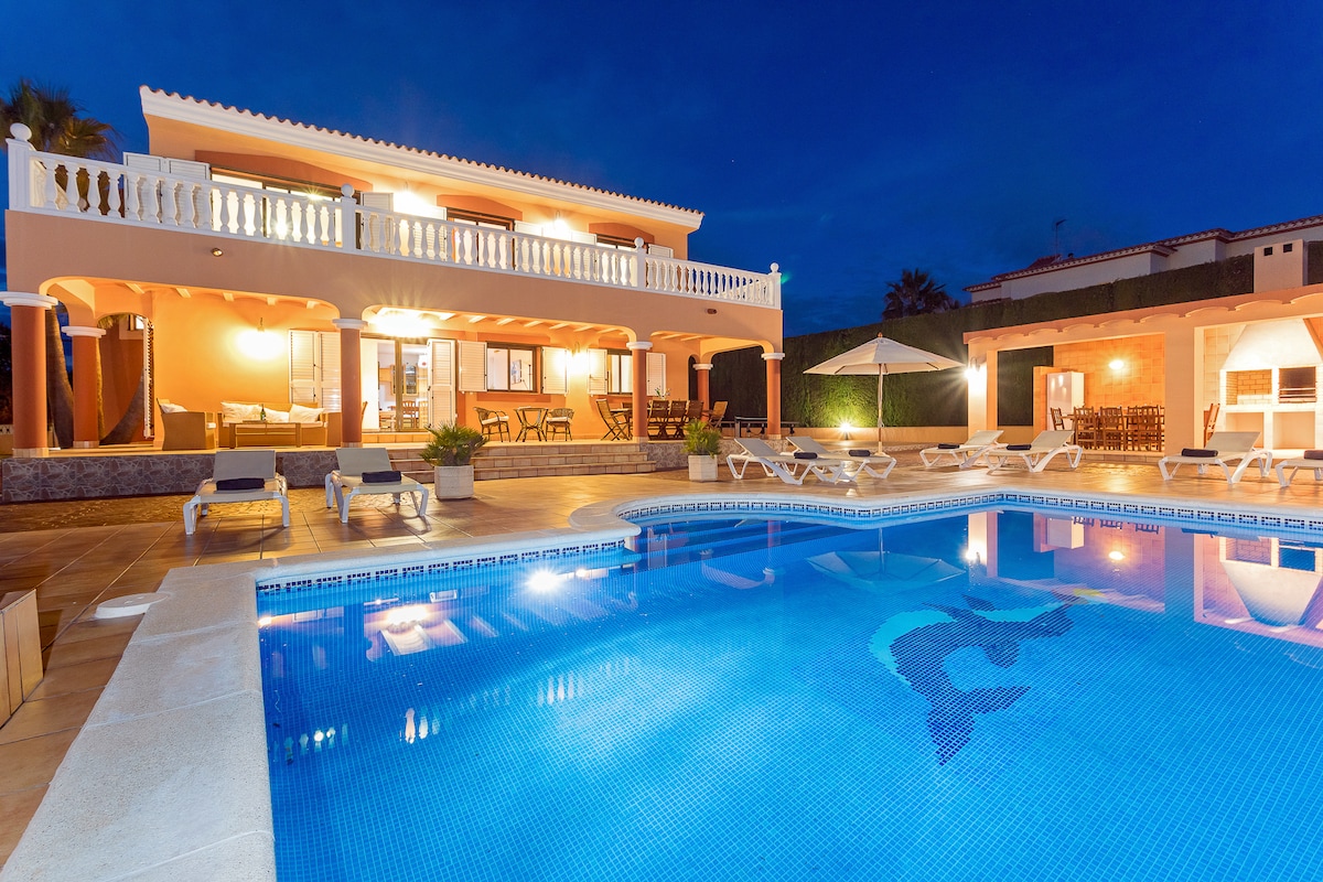 The villa exterior glows warmly at night, with illuminated windows and patio areas. A spacious swimming pool features a dolphin design at its center, surrounded by cushioned sun beds and stylish outdoor dining spaces, creating a serene atmosphere for relaxation.