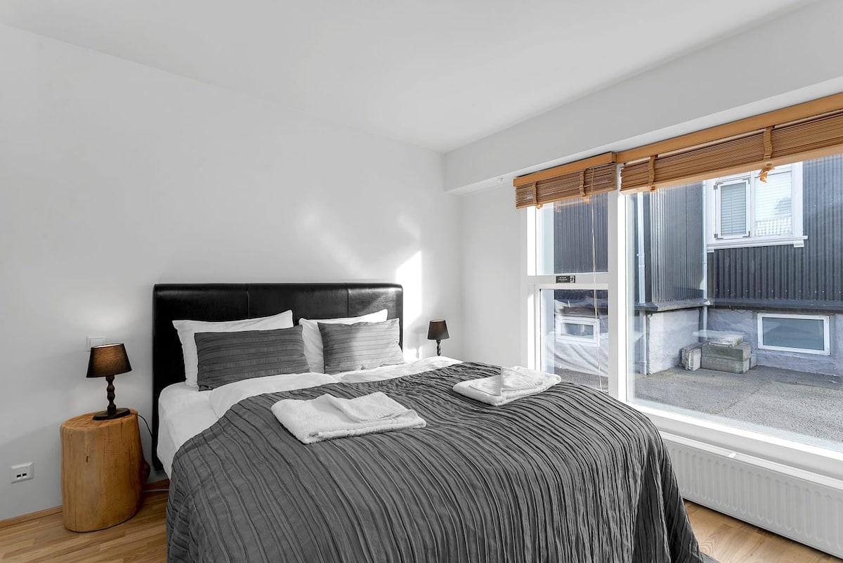 A bright and airy bedroom features a spacious bed with an elegant grey coverlet. Two bedside lamps are positioned on either side, and sunlight streams in through a large window adorned with wooden blinds, offering a view of the exterior.