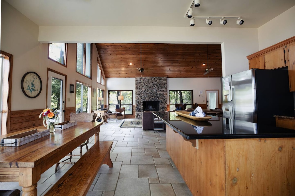 An open floor plan is highlighted, featuring a spacious kitchen with a dark countertop and wooden cabinetry. High vaulted ceilings are seen above a rustic dining table, complemented by stone accents around a large fireplace. Large windows allow natural light to brighten the living area.