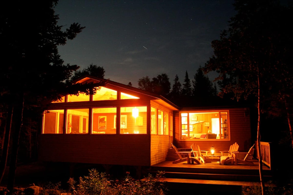 The cottage is softly illuminated at night, highlighting its modern architecture and expansive windows. A cozy seating area is visible inside, while a firepit and wooden Muskoka chairs are placed outside on the porch, surrounded by trees and a starry sky.