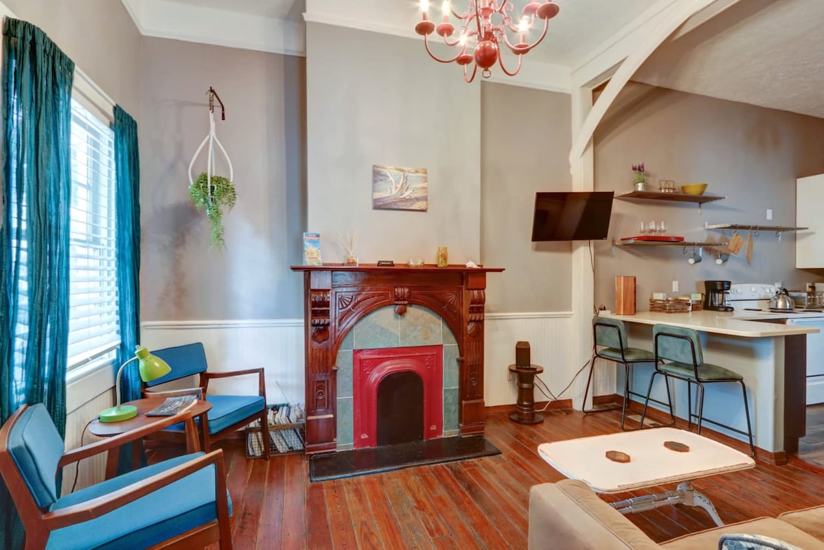 A cozy living area features hardwood floors and a decorative fireplace with a bold red surround. Modern seating is arranged for comfort, complemented by a chandelier overhead. Natural light filters through teal curtains, enhancing the space's inviting ambiance while a television is mounted on the wall.
