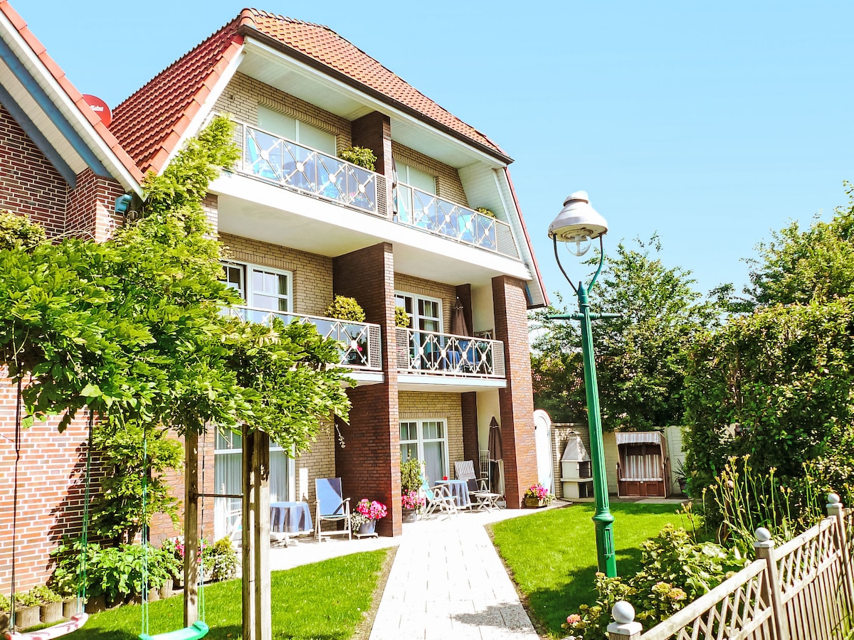 The exterior of a three-story apartment building showcases a brick facade, with balconies adorned with potted plants and chairs. A garden path leads through a manicured lawn, bordered by colorful flowers and greenery, inviting guests to enjoy the outdoor space.