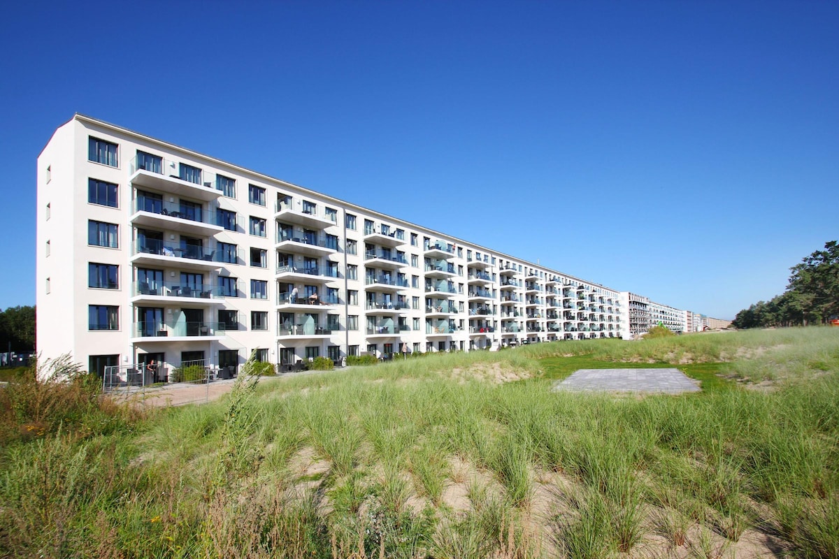 A modern multi-story building stretches along the landscape, showcasing numerous balconies. Green grass and natural vegetation are observed in the foreground, creating a connection to the surrounding environment. The clear blue sky provides a bright backdrop to the structure, enhancing the overall setting.