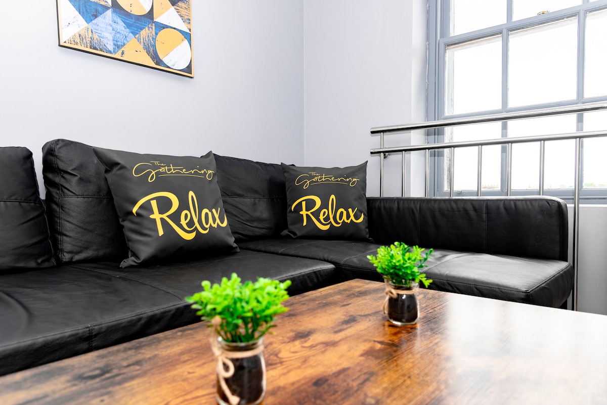 A modern lounge area features a black L-shaped sofa adorned with two decorative pillows that read 'Gathering' and 'Relax.' A wooden coffee table with small potted plants sits in the center, creating a welcoming space for relaxation.
