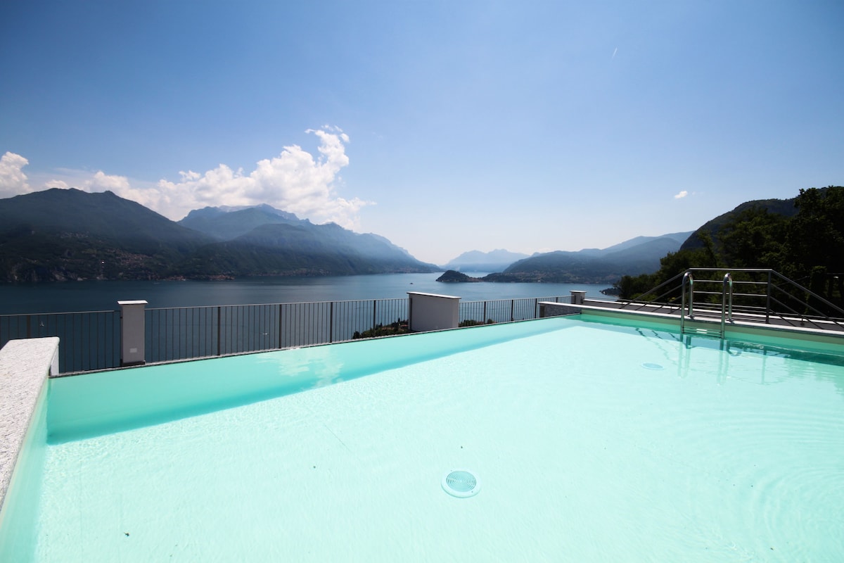 A roof top swimming pool is shown with clear turquoise water, reflecting the blue sky. Expansive views of the lake and surrounding mountains create a serene backdrop, enhancing the tranquil atmosphere. The pool area features a surrounding railing that provides safety and unobstructed sightlines.