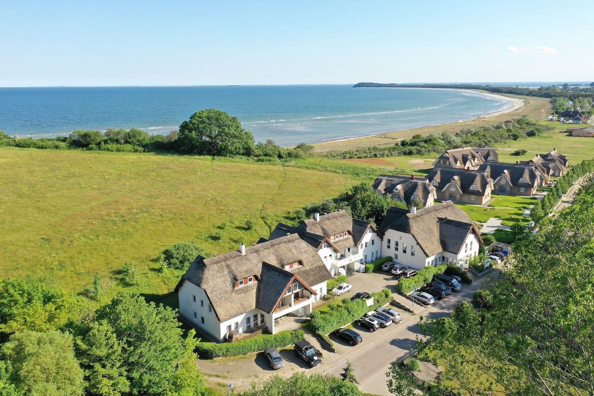 Aerial view of a scenic coastal area featuring a cluster of thatched-roof buildings, surrounded by lush greenery. The nearby sandy beach curves along the coastline, stretching into the horizon, with the tranquil sea visible in the background.