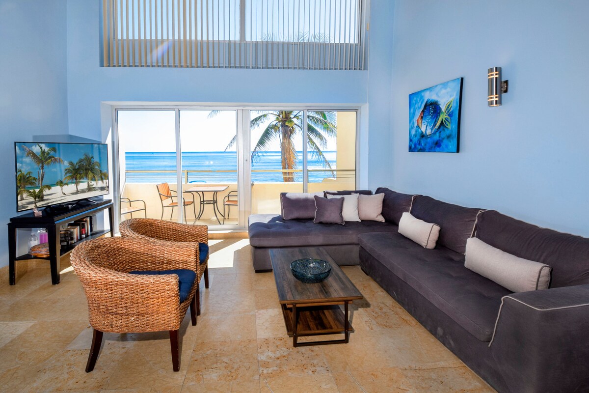 A spacious living area is highlighted by a large sectional sofa complemented by decorative cushions. A television sits on a stand near a pair of rattan chairs. Floor-to-ceiling windows offer views of the ocean and patio area, allowing natural light to fill the room.