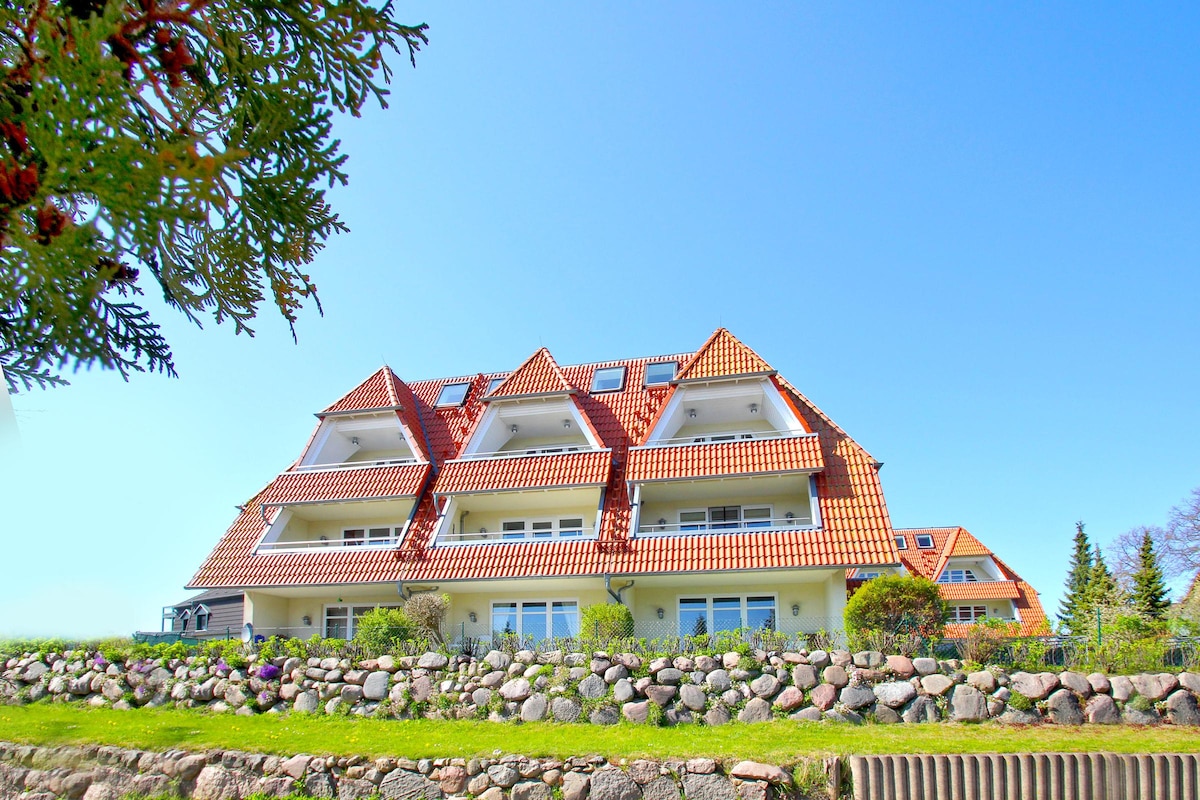 A well-maintained building featuring multiple balconies under a clear blue sky. The structure is complemented by a stone wall with greenery, enhancing the inviting appearance of the property.