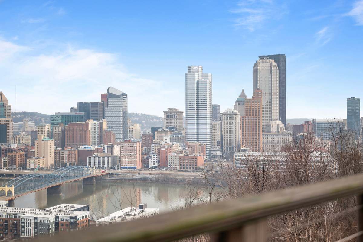 A panoramic view of downtown Pittsburgh is captured, featuring a skyline of modern buildings and bridges over the river. The foreground includes trees and natural scenery, creating a contrast with the urban landscape in the background under a clear blue sky.
