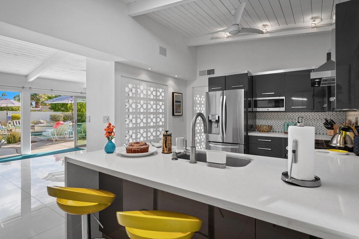 A modern kitchen showcases sleek grey cabinetry and stainless steel appliances. Bright yellow bar stools are positioned at a white quartz island, which holds decorative items including a flower vase and a cake. Natural light floods in through large windows, highlighting the open-concept design.