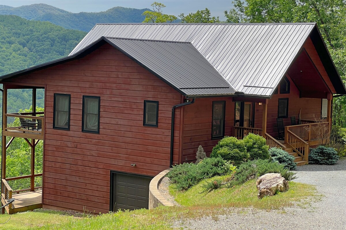 The cabin's exterior is crafted from rich wood, complemented by a dark metal roof. Surrounded by lush greenery and mountains, it features a welcoming entryway with a spacious porch and steps leading to the front door. Large windows enhance natural light and views.